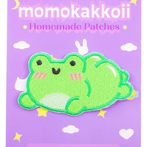 Könnte beinhalten: Grüner Filz-Frosch-Patch mit einem rosafarbenen Lächeln und Rouge. Der Patch befindet sich auf einem lila Hintergrund mit weißen Wolken und Sternen. Der Text "momokakkoii.com" befindet sich am unteren Rand des Bildes.
