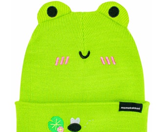 Puede incluir: Un gorro de punto verde brillante con un diseño de cara de rana. El gorro presenta detalles bordados que incluyen una boca sonriente, mejillas rosas y ojos de rana. El borde del gorro tiene un pequeño diseño bordado y una etiqueta negra con el texto "momokakkoii".