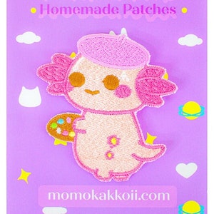 May include: Pink axolotl wearing a pink beret and holding a paint palette. The axolotl is surrounded by stars, clouds, and planets. The text "momokakkoii" and "Homemade Patches" is at the top of the image. The text "momokakkoii.com" is at the bottom of the image.