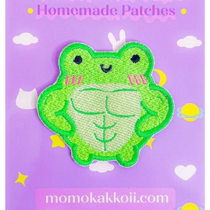 Puede incluir: Parche verde bordado para planchar de una rana de dibujos animados con músculos abultados. La rana sonríe y tiene las mejillas rosas. El parche está sobre un fondo morado con nubes blancas y estrellas. El texto "momokakkoii.com" está en la parte inferior de la imagen.