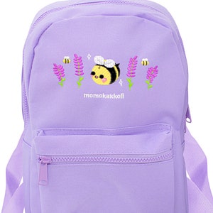 Könnte beinhalten: Ein lilafarbener Rucksack mit einer niedlichen gestickten Biene und Blumen. Der Rucksack hat eine Reißverschlusstasche auf der Vorderseite und verstellbare Träger. Der Text "momokakkoii" ist auf der Vorderseite des Rucksacks gestickt.