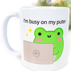May include: White ceramic mug with a green frog cartoon character using a laptop. The mug has the text "I'm busy on my puter" printed on it.