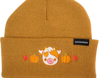 Puede incluir: Un gorro de punto marrón con una vaca y calabazas bordadas. El gorro tiene una etiqueta negra con el texto "momokakkoi" en el lateral.