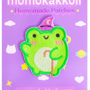 Könnte beinhalten: Ein grüner Frosch, der einen rosa Zaubererhut mit einer Sichelmondspitze trägt. Der Frosch hält einen braunen Spazierstock. Der Frosch ist auf einem weißen Patch mit violettem Hintergrund gestickt. Der Text "momokakkoii.com" befindet sich am unteren Rand des Patches.