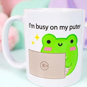 Può includere: Tazza in ceramica bianca con un'illustrazione di una rana verde che utilizza un laptop. La tazza ha il testo "I'm busy on my puter" in nero. La rana ha le guance rosa ed è circondata da stelle gialle. La tazza ha un manico bianco.