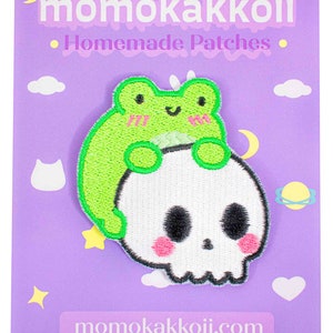 May include: A green frog with pink cheeks sits on top of a white skull with pink cheeks. The image is on a purple background with white clouds and stars. The text "momokakkoii.com" is at the bottom of the image.
