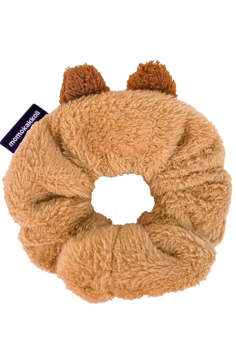 K&ouml;nnte beinhalten: Ein braunes, flauschiges Haargummi mit zwei braunen, flauschigen B&auml;renohren oben. Das Haargummi hat ein violettes Etikett mit dem Text "momokakkoi" darauf.