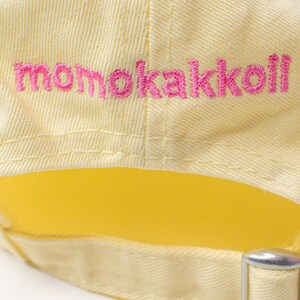 以下が含まれることがあります： ピンクの刺繍ロゴが「momokakkoli」と入った黄色のベースボールキャップ