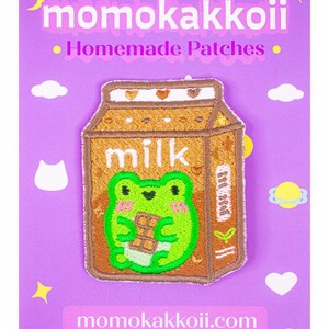 May include: Embroidered patch featuring a cartoon frog holding a chocolate bar inside a milk carton. The patch has the text "milk" and "momokakkoii.com" on a purple background with decorative elements. The patch is a homemade product.