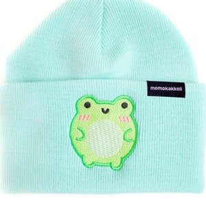 May include: A light blue knit beanie with a green embroidered frog on the front. The beanie has a black label with the text "momokakkoi" on the side.
