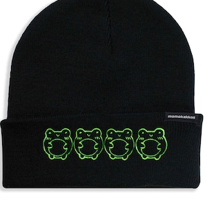 May include: Black knit beanie with a green embroidered design of four frogs hugging each other. The beanie has a small black label with the text "momokakkoii" on the side.