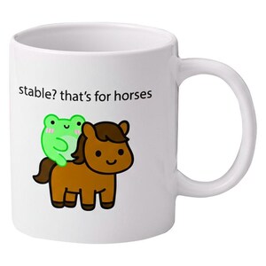 Op de afbeelding: Witte keramische mok met een cartoon illustratie van een bruin paard met een groene kikker op zijn rug. De mok heeft de tekst "stable? that's for horses" erop. De mok heeft een wit handvat.