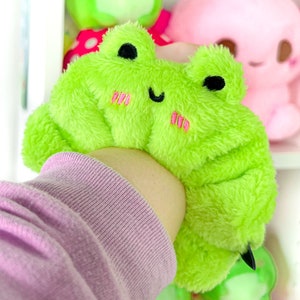 May include: A bright green plush frog with a smiling face and pink blush. The frog is wearing a pink sweater and has a fluffy texture.