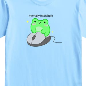 May include: Light blue t-shirt featuring a cartoon frog sitting on a computer mouse. The frog is green with pink cheeks and the text "mentally elsewhere" is above the frog. A small pink square with Japanese text is on the upper left.
