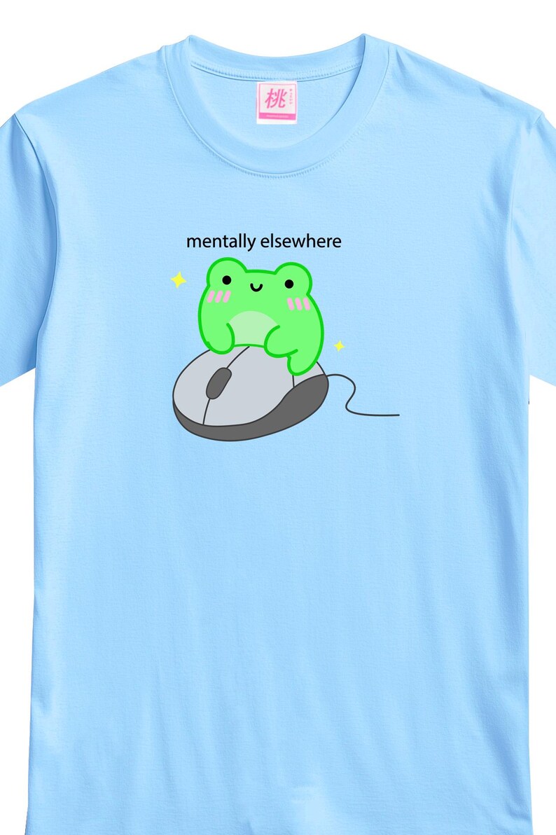 May include: Light blue t-shirt featuring a cartoon frog sitting on a computer mouse. The frog is green with pink cheeks and the text "mentally elsewhere" is above the frog. A small pink square with Japanese text is on the upper left.