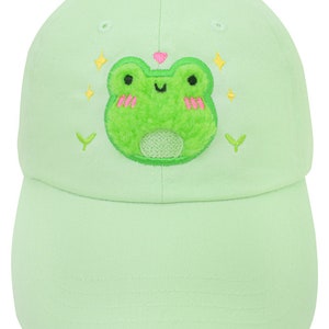 Könnte beinhalten: Eine hellgrüne Baseballkappe mit einem grünen Frosch, der auf der Vorderseite gestickt ist. Der Frosch hat rosa Wangen und ein rosa Herz darüber. Um den Frosch herum befinden sich gelbe Sterne und grüne Blätter.