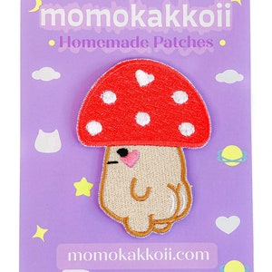 May include: A red and white mushroom with a cute face and a pink heart on the cap. The mushroom is embroidered on a light purple background with the text "momokakkoii.com" and "Homemade Patches".