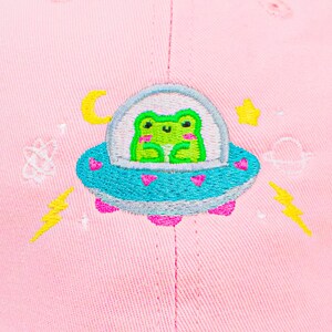 Puede incluir: Gorra de b&eacute;isbol rosa con un dise&ntilde;o bordado de una rana verde en un platillo volante con corazones, estrellas y rayos.