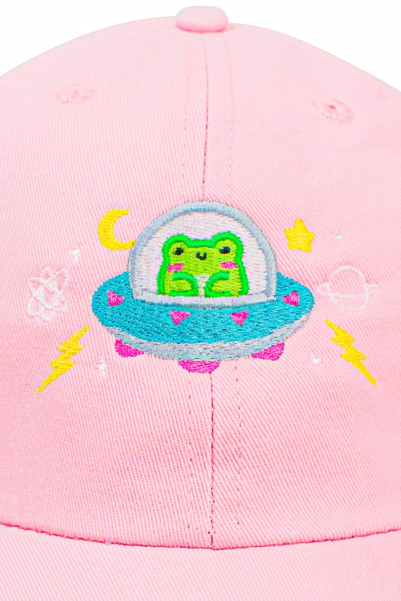 Puede incluir: Gorra de b&eacute;isbol rosa con un dise&ntilde;o bordado de una rana verde en un platillo volante con corazones, estrellas y rayos.