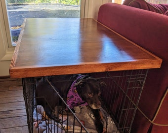 May include: A brown wooden table top with a black metal dog crate underneath. A brown and black dog is inside the crate, lying on a gray and pink blanket. The dog is wearing a purple and black bandana with a Halloween-themed design.