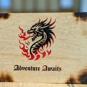 May include: A wooden tabletop gaming screen with a dragon and wing design. The screen is made of light-colored wood with dark burnt edges and metal corner protectors. The text "Adventure Awaits" is below the dragon graphic.