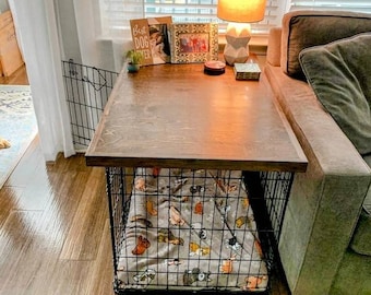 May include: A wooden table top with a black metal dog crate underneath. The crate is lined with a patterned blanket. The table is next to a couch with a lamp on it. The text on the card on the table reads "Best Dog Ever".