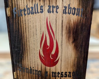 May include: Wooden sign with a charred border and the text "Fireballs are about sending a message." A red flame graphic is centered on the light-colored wood. The sign is hinged with a small brass hinge.