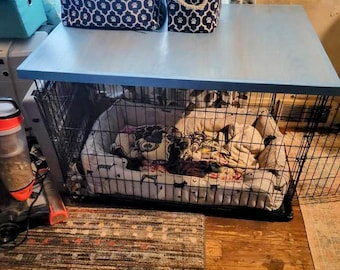 May include: A blue painted wooden table top with two blue and white storage bins on top. The table top is over a black metal dog crate with a black and white patterned dog bed inside.