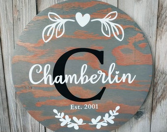 May include: A round wooden sign with a distressed gray and brown paint finish. The sign features a large black letter "C" and the name "Chamberlin" in white cursive lettering. The sign also includes a white heart and floral designs, and the text "Est. 2001".