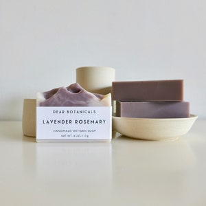 Pu&ograve; includere: Barrette di sapone artigianali con lavanda e rosmarino. Una barretta ha un design marmorizzato viola e bianco, con un'etichetta che recita "Dear Botanicals Lavender Rosemary". Due barrette aggiuntive sono impilate su un piccolo piatto bianco sporco. Il sapone &egrave; di 113 g.
