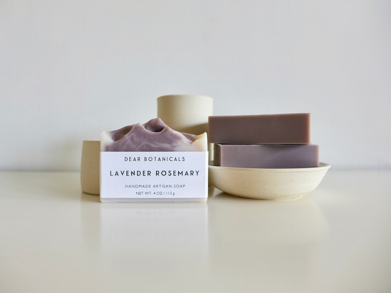Pu&ograve; includere: Barrette di sapone artigianali con lavanda e rosmarino. Una barretta ha un design marmorizzato viola e bianco, con un'etichetta che recita "Dear Botanicals Lavender Rosemary". Due barrette aggiuntive sono impilate su un piccolo piatto bianco sporco. Il sapone &egrave; di 113 g.