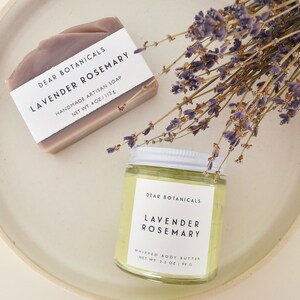 Pu&ograve; includere: Una saponetta artigianale alla lavanda e rosmarino e un vasetto di burro corpo montato, entrambi etichettati "Dear Botanicals Lavender Rosemary", sono esposti con rametti di lavanda essiccata. Il sapone pesa 113 g, e il burro corpo pesa 90 g.