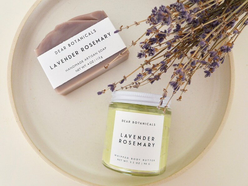 Pu&ograve; includere: Una saponetta artigianale alla lavanda e rosmarino e un vasetto di burro corpo montato, entrambi etichettati "Dear Botanicals Lavender Rosemary", sono esposti con rametti di lavanda essiccata. Il sapone pesa 113 g, e il burro corpo pesa 90 g.