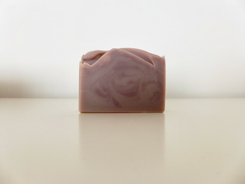 Pu&ograve; includere: Una saponetta rettangolare fatta a mano, di colore viola chiaro, con un motivo a spirale. Il bordo superiore ha un taglio decorativo e irregolare. Il sapone &egrave; appoggiato su una superficie chiara, con uno sfondo bianco.