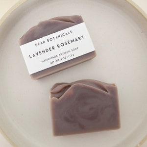 Pu&ograve; includere: Due saponi artigianali alla lavanda e rosmarino su un piatto di ceramica bianco. I saponi sono di colore viola tenue con una consistenza a spirale. Ogni sapone ha un'etichetta bianca che recita "DEAR BOTANICALS LAVENDER ROSEMARY HANDMADE ARTISAN SOAP NET WT. 4 OZ / 113g."