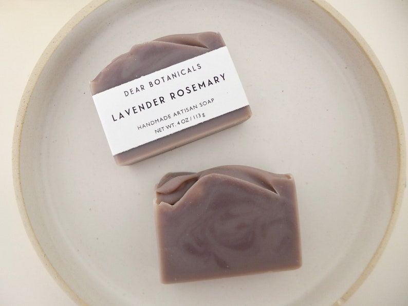 Pu&ograve; includere: Due saponi artigianali alla lavanda e rosmarino su un piatto di ceramica bianco. I saponi sono di colore viola tenue con una consistenza a spirale. Ogni sapone ha un'etichetta bianca che recita "DEAR BOTANICALS LAVENDER ROSEMARY HANDMADE ARTISAN SOAP NET WT. 4 OZ / 113g."