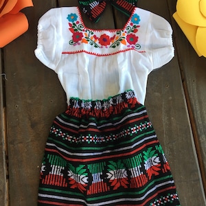 May include: A white embroidered blouse with a black and green skirt with white lace trim. The skirt has a red, white, and green pattern. A black and white bow with red and blue accents is included.