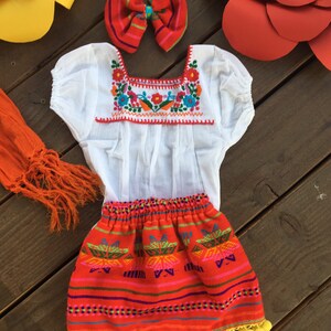 May include: A white embroidered blouse with a red and white trim, a red skirt with a colorful geometric pattern, and a red bow with a pink and white stripe. The skirt has a yellow pom-pom trim.