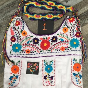 May include: White tote bag with colorful floral embroidery. The bag has a purple trim and a rainbow woven strap. The bag has a label that says "Maridla Artesanal".