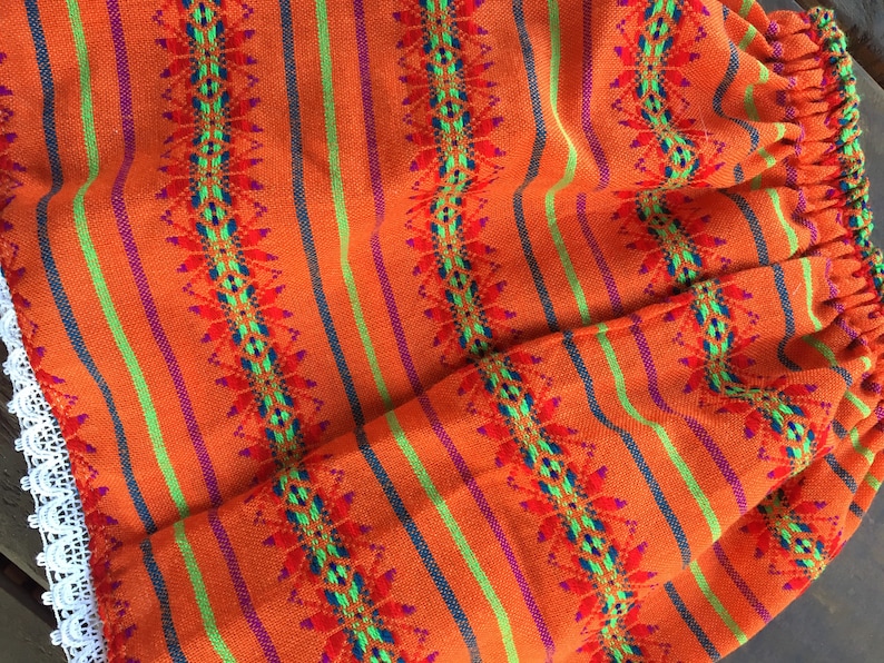 May include: Orange, green, and purple striped fabric with a geometric pattern. The fabric has a white lace trim.