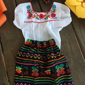 May include: A white embroidered blouse with short sleeves and a black skirt with colorful embroidery and white lace trim. A black and colorful bow headband is included.