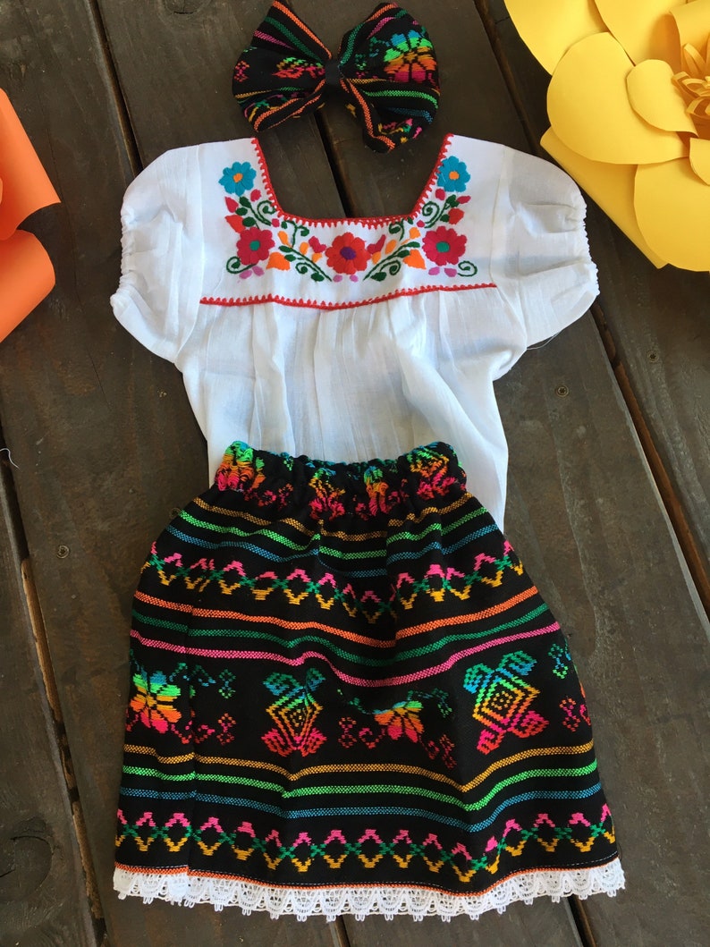 May include: A white embroidered blouse with short sleeves and a black skirt with colorful embroidery and white lace trim. A black and colorful bow headband is included.