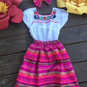 May include: A white embroidered blouse with a pink and multi-colored striped skirt. The skirt has a white lace trim. A pink and white bow is included.