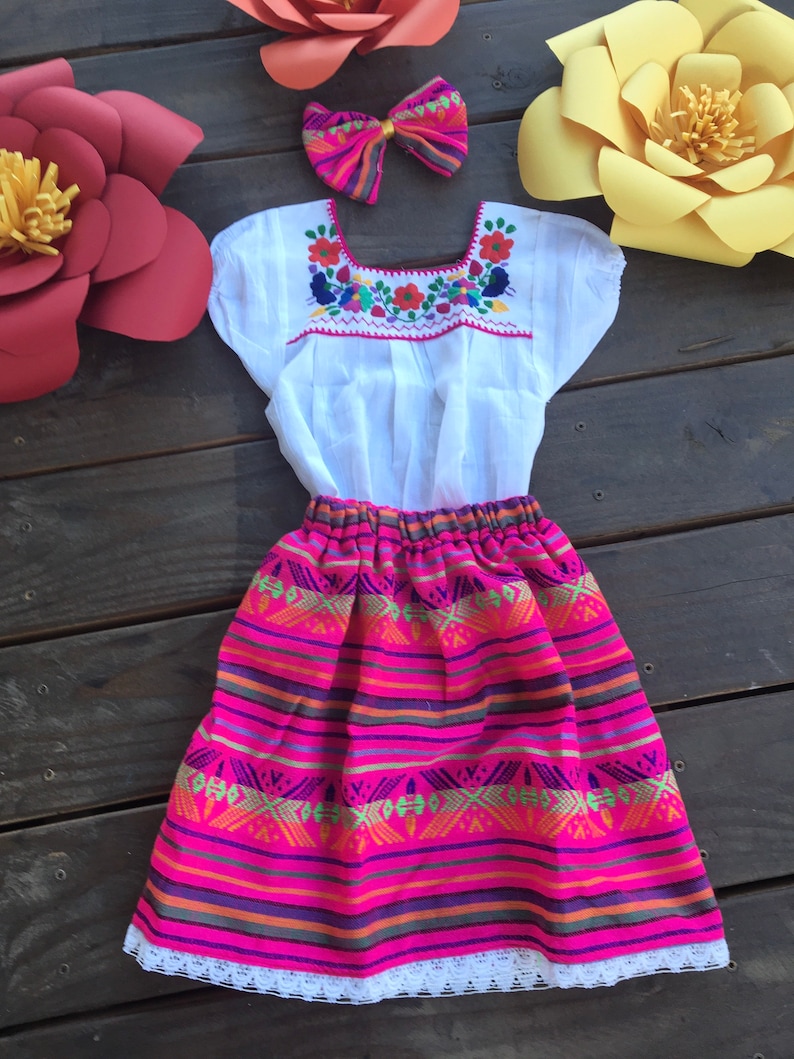 May include: A white embroidered blouse with a pink and multi-colored striped skirt. The skirt has a white lace trim. A pink and white bow is included.