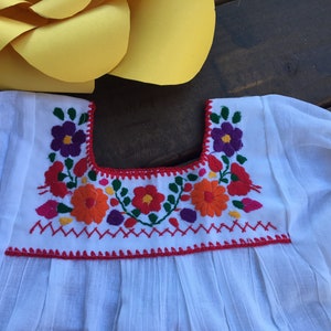 May include: White cotton blouse with a red embroidered neckline featuring colorful floral designs.
