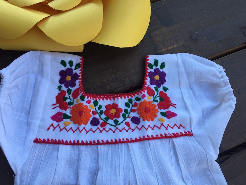 May include: White cotton blouse with a red embroidered neckline featuring colorful floral designs.