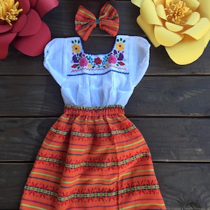 May include: A white embroidered blouse with a bright orange skirt and a red and white bow. The skirt has a white lace trim. The outfit is a traditional Mexican style.