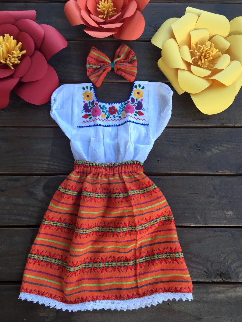 May include: A white embroidered blouse with a bright orange skirt and a red and white bow. The skirt has a white lace trim. The outfit is a traditional Mexican style.