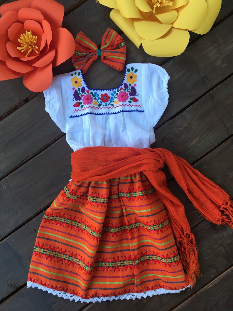 May include: A white embroidered blouse with a bright orange skirt and matching shawl. The skirt has a white lace trim and the blouse has a colorful floral design. The outfit is perfect for a fiesta or celebration.