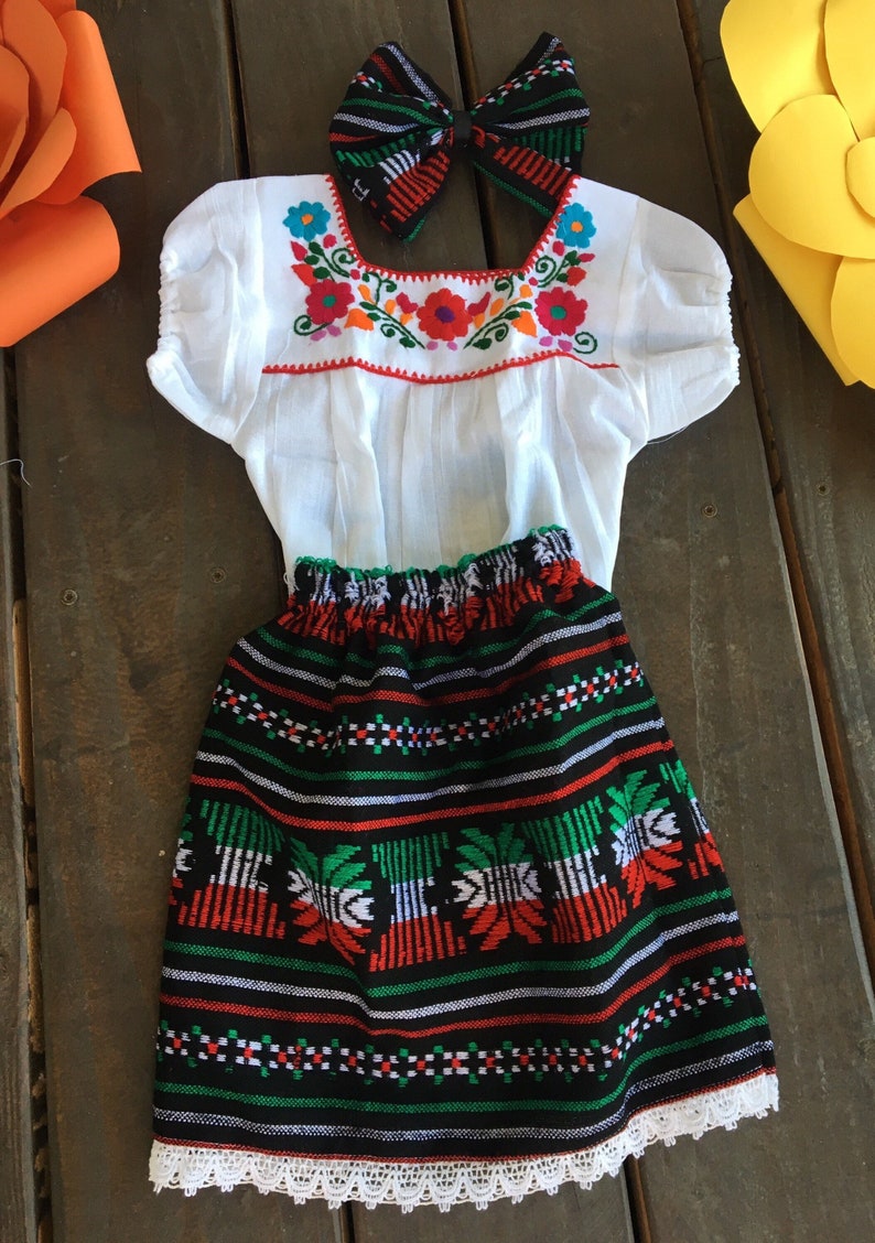 May include: A white embroidered blouse with a black and green skirt with white lace trim. The skirt has a red, white, and green pattern. A black and white bow with red and blue accents is included.